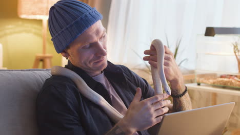 close up view of man sitting on a sofa holding a snake while using the laptop at home