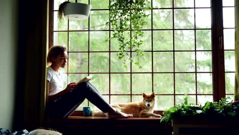 attractive young lady is reading book sitting on windowsill in the house together with adorable puppy. large window, green plants, nice interior is visible.