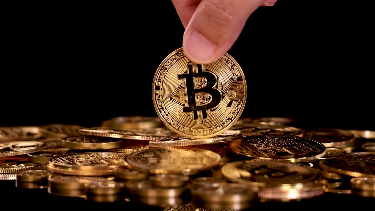 Hand releases gold Bitcoin coin onto pile of coins, dramatic lighting, black background, close-up