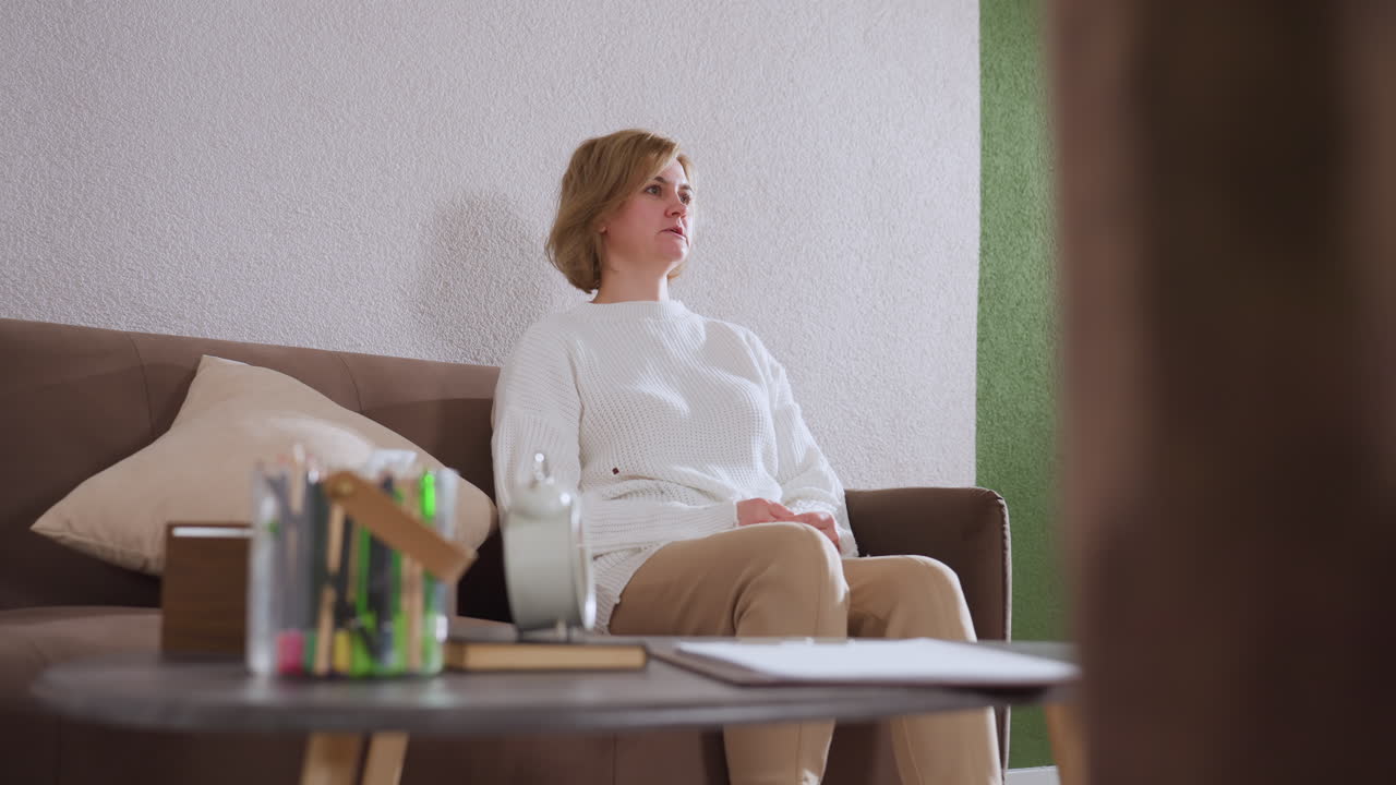 Mindful guest seated on sofa engaged in conversation demonstrating with hands, pen pack visible on table, neutral wall and green accent wall background, soft lighting evokes calm therapy environment