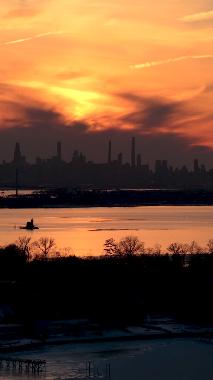 Dramatic orange sky with dark clouds during a winter sunset, silhouetting the iconic new york city skyline as seen across a frozen bay with snowy shores and silhouetted trees in the foreground