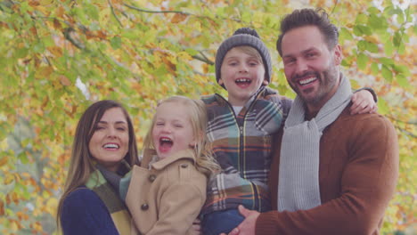 Portrait-Of-Family-With-Mature-Parents-Carrying-Two-Children-In-Front-Of-Autumn-Leaves