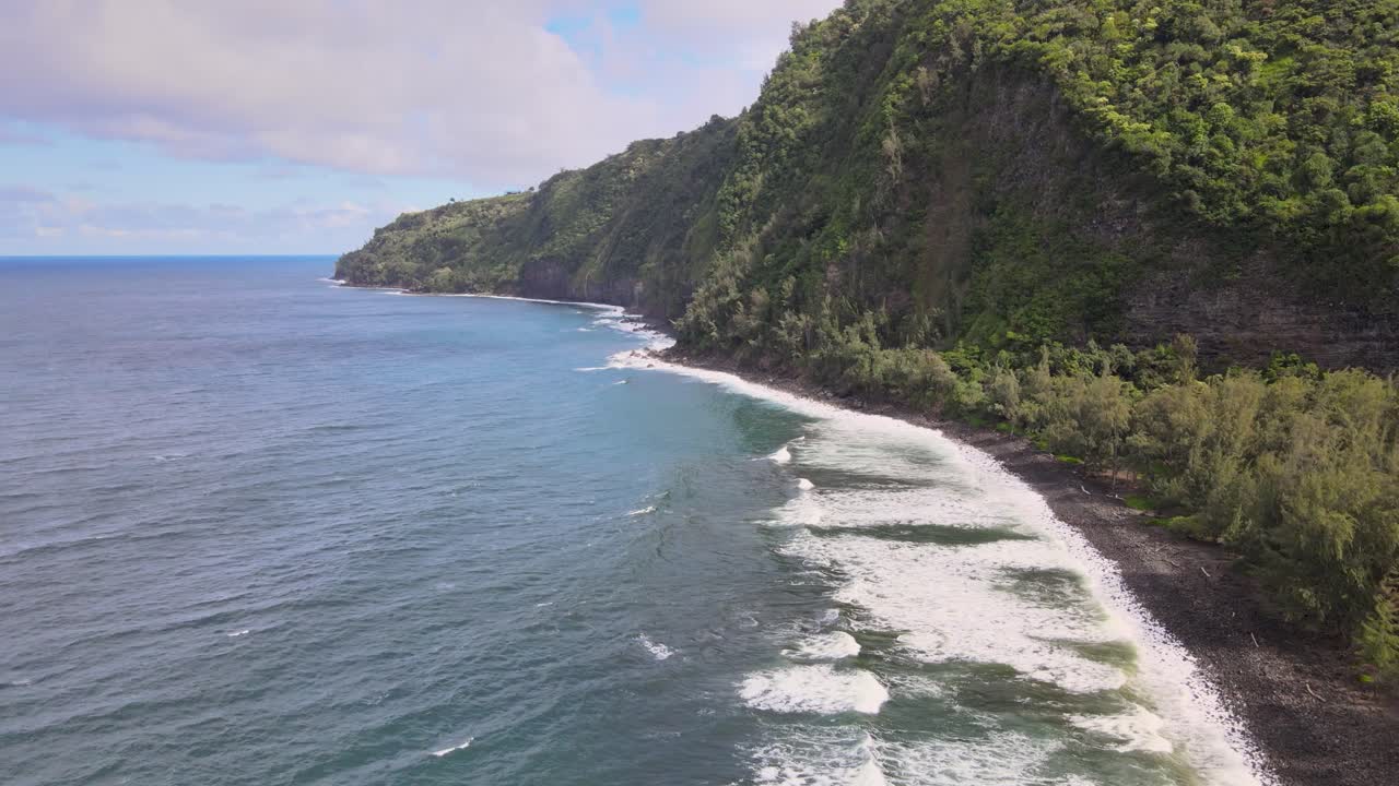Drone capture revealing the scenic side of Waipi'o Valley from a black sand beach under partial sunlight, with moderate waves on Hawaii's Big Island