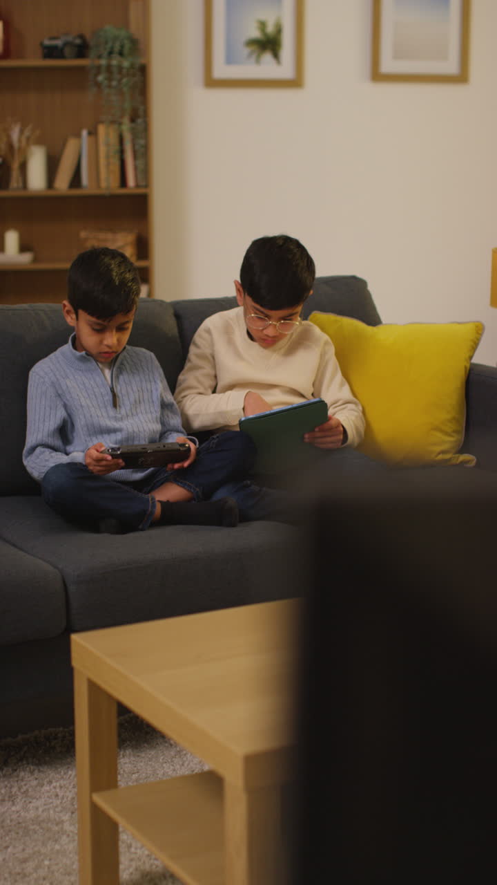 Vertical Video Of Two Young Boys Sitting On Sofa At Home Playing Games Or Streaming Onto Digital Tablet And Handheld Gaming Device