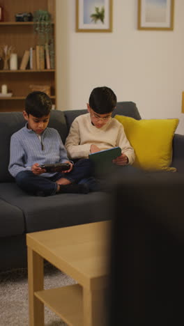 vertical video of two young boys sitting on sofa at home playing games or streaming onto digital tablet and handheld gaming device