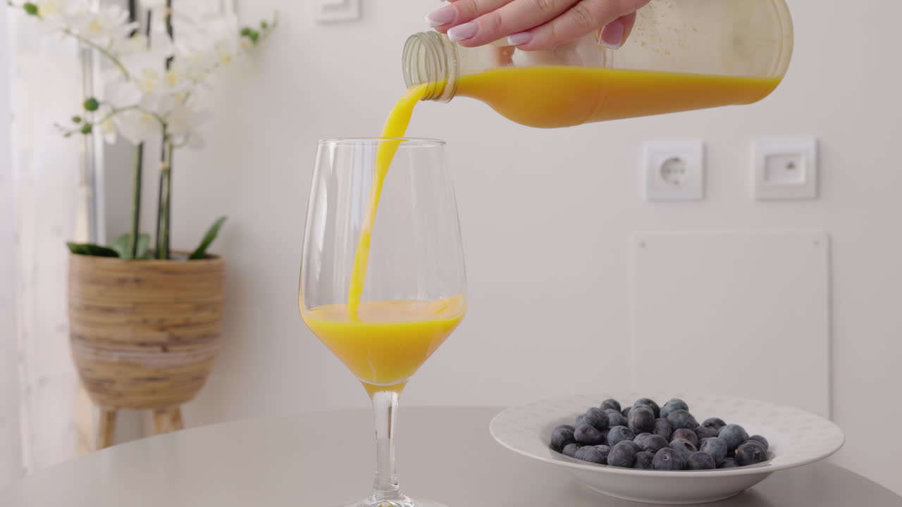 Mano che versa succo d'arancia fresco in un bicchiere sul tavolo della cucina, mirtilli su un piatto in un moderno interno domestico luminoso.