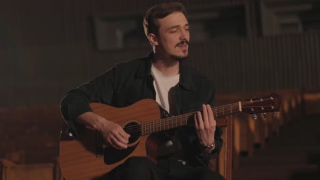 a handsome guy plays an acoustic guitar in an abandoned cinema. the musician sings a song and accompanies on the guitar