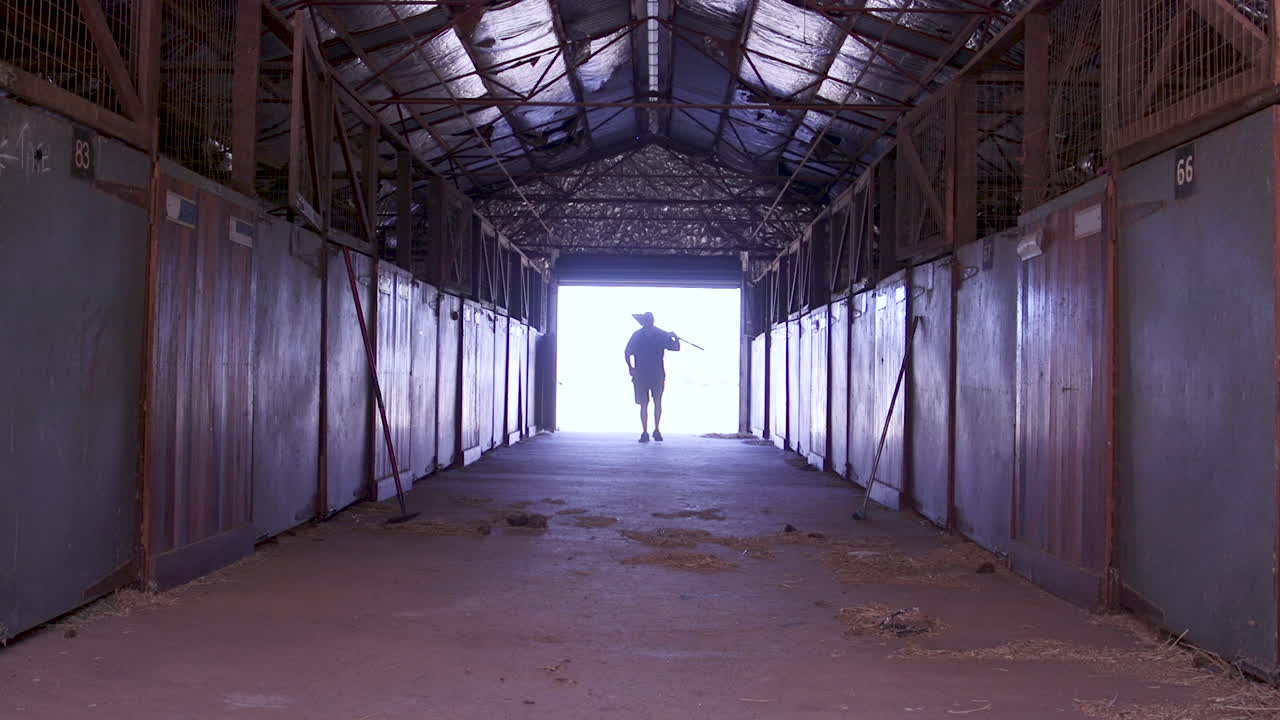 A Stable hand worker carrying a shovel over his shoulder through the horse's stables