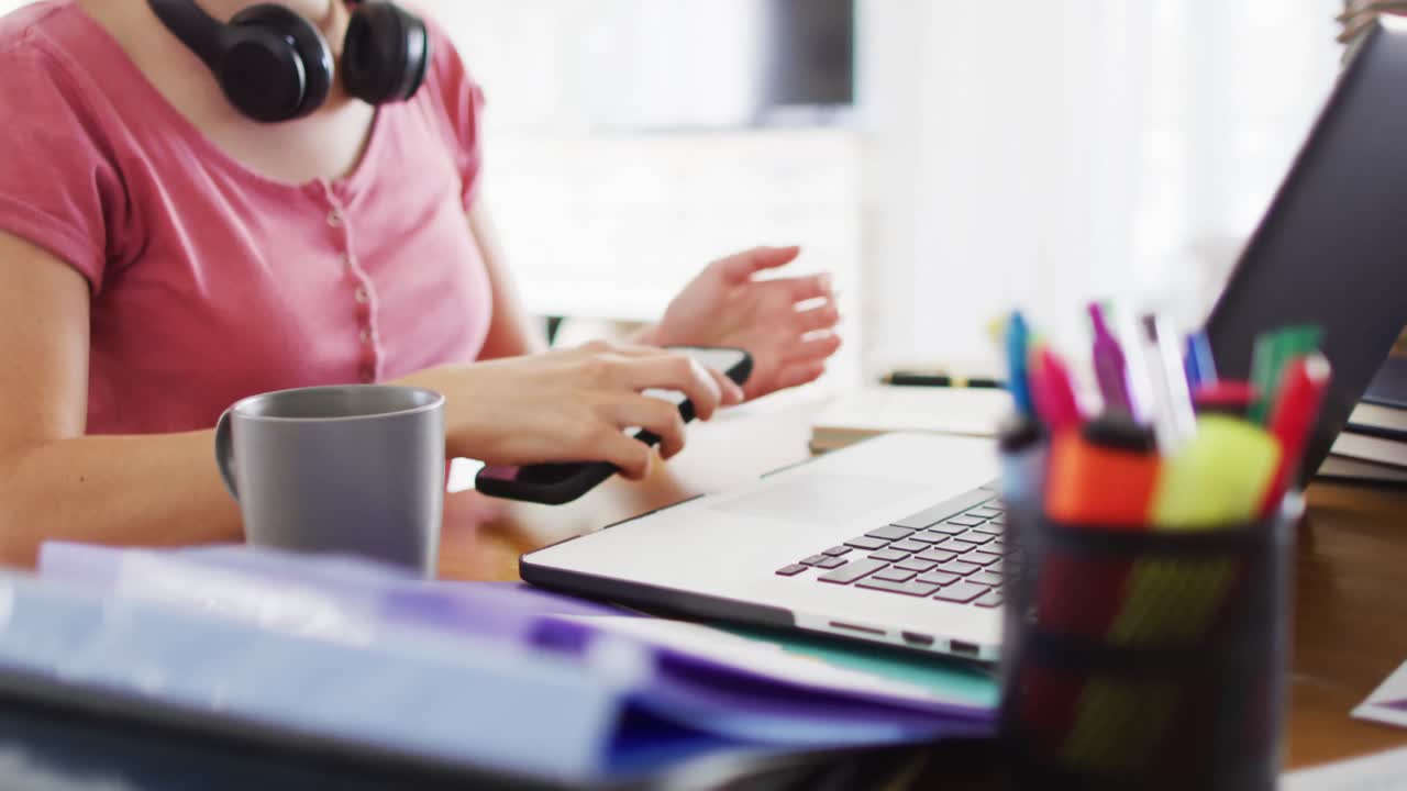 mujer caucásica sentada en un escritorio, enviando mensajes de texto en un teléfono inteligente y usando una computadora portátil