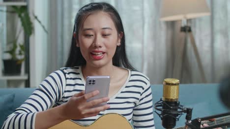 close up of asian woman reading comments on smartphone and speaking to camera while live stream playing guitar at home