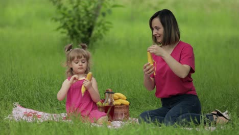 Family-weekend-at-picnic.-Daughter-child-girl-with-mother-on-grass-meadow-eating-bananas,-having-fun