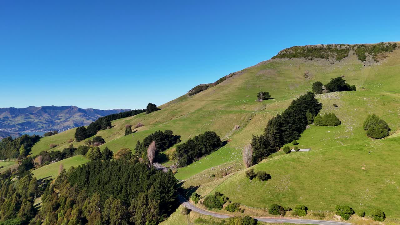 Aerial footage captures rolling hills, lush greenery, and a serene lake under clear blue skies in Akaroa, New Zealand