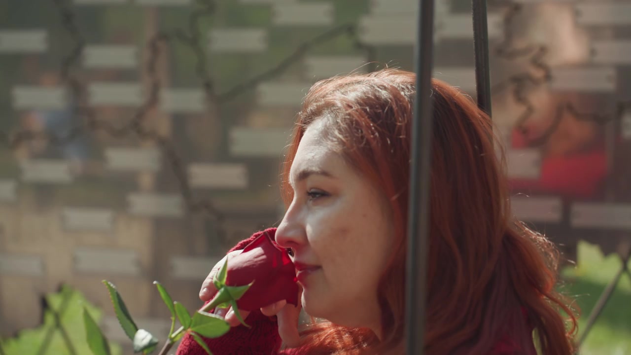 Primo piano di donna con capelli rossi che tiene in mano e annusa una singola rosa mentre è seduta su un'altalena all'aperto. La donna indossa un maglione a maglia testurizzata, la sua espressione è calma mentre interagisce delicatamente con il fiore.