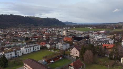 Swiss-town-with-buildings-and-rolling-hills-in-the-backdrop,-cloudy-day,-aerial-view