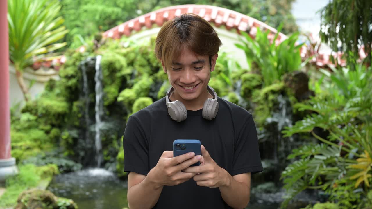 Young Thai Asian man in traditional Asian Zen garden during summer using phone. Confident, relaxed, and authentic look suitable for various concepts and uses