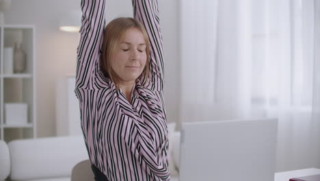 tired student woman is learning in home sitting at table and writing stretching hands up relaxing back