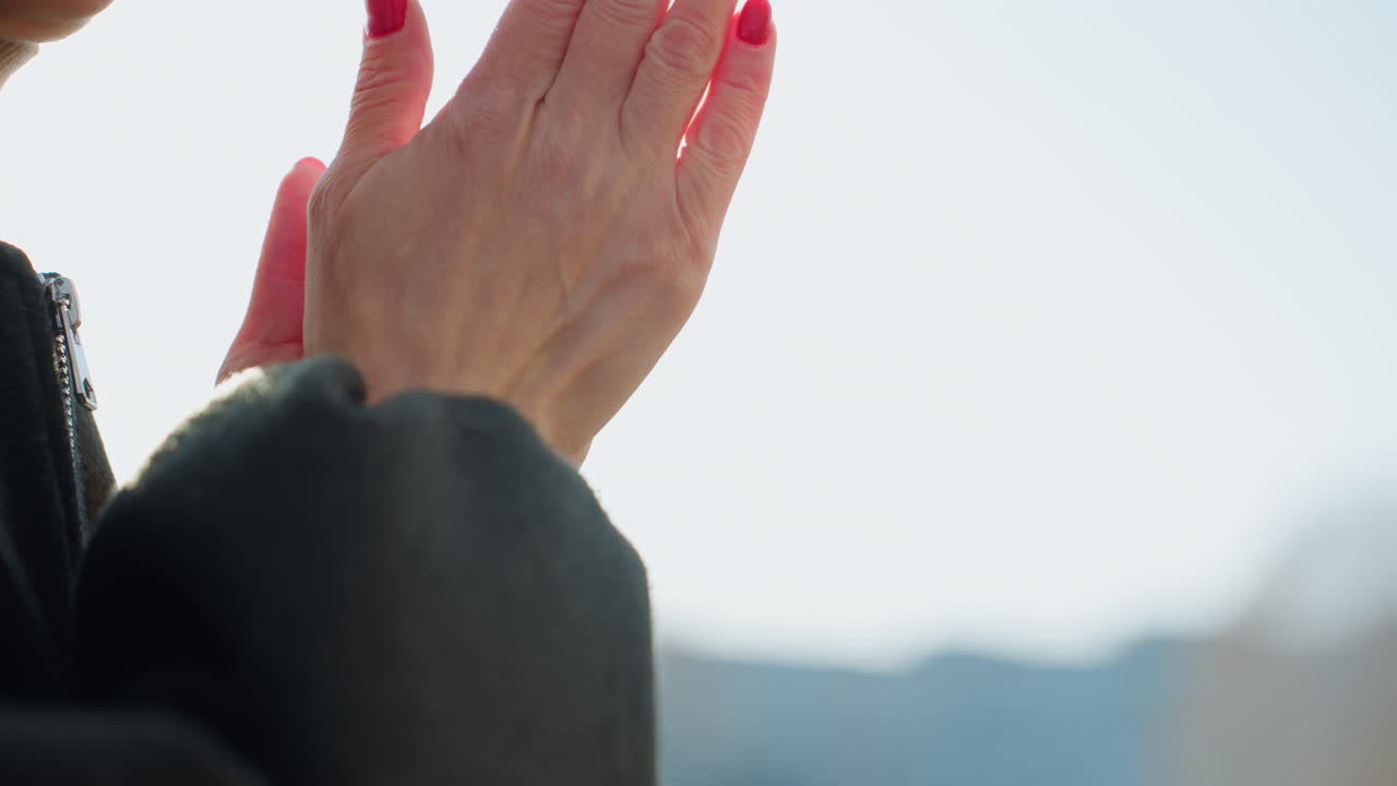 Close up of mature woman with red manicure covering mouth with hands under sunlight, warm golden rays shining through fingers, showing happiness, gratitude in outdoor setting