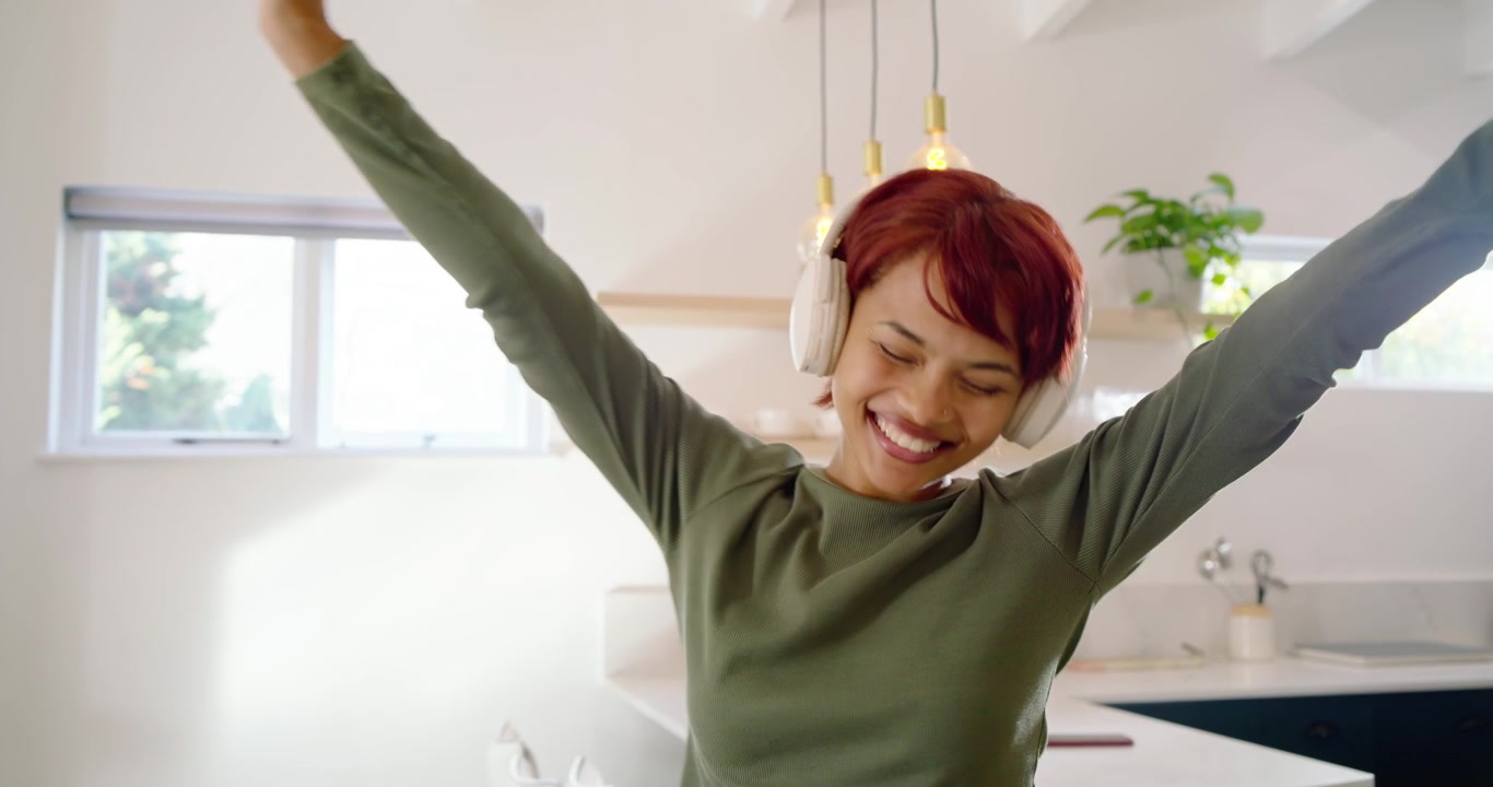 Adult African American woman hearing music, moving, adjusting headphones in kitchen wearing green