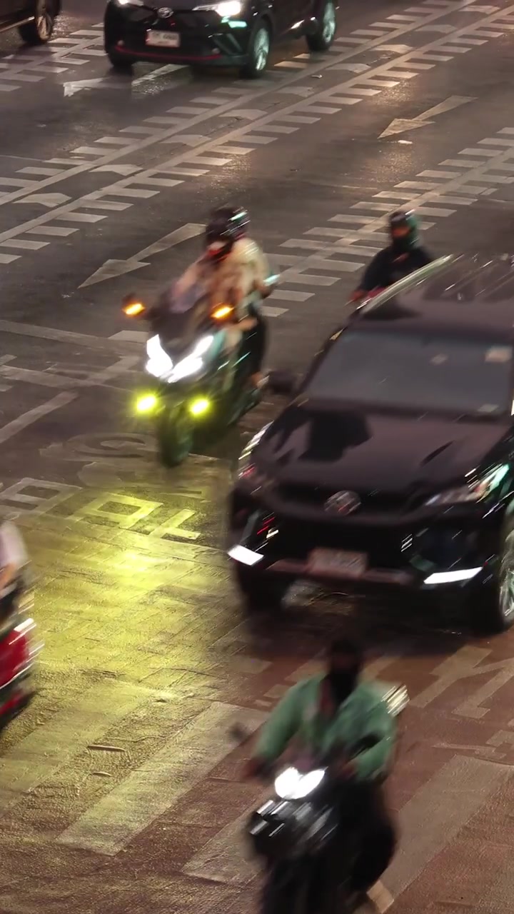 A high-angle vertical shot showing cars, taxis, and motorcycles navigating a busy urban intersection.