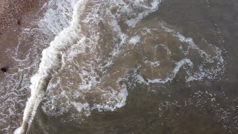 Swirling-waves-in-murky-North-Sea-crashing-on-sandy-beach-covered-in-seaweed