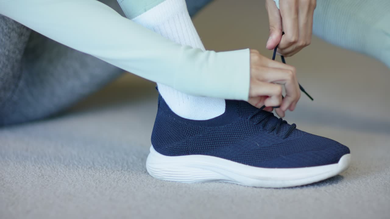 Donna afroamericana che tira i lacci blu scuro e allaccia la sneaker sul tappeto, in leggings, preparandosi ad allenarsi.