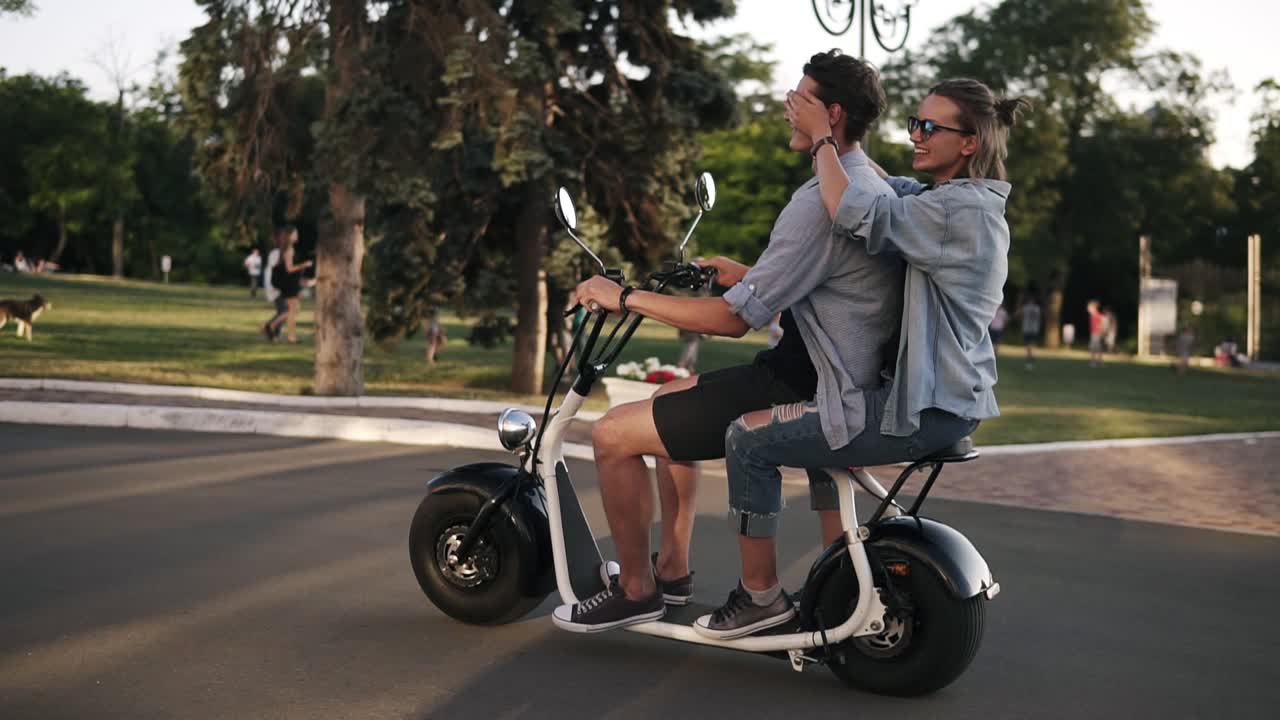 Young couple on dating riding an electronic minibike on the street. Wearing casual. Blonde lady closes his boyfriend's eyes while riding. Daytime