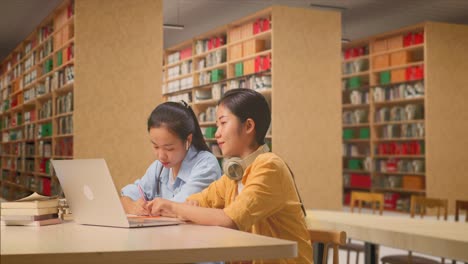 asian woman student teaching her classmate on a laptop while sitting on a table in the library