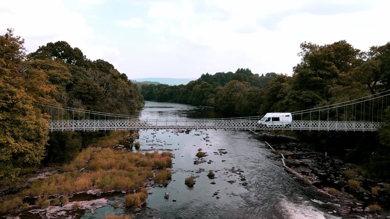 A white panel van drives over a steel suspension bridge spanning over a river with lush trees on either bank of the river in Wales United Kingdom
