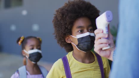 diverse male teacher measuring temperature of schoolchildren, all wearing face masks