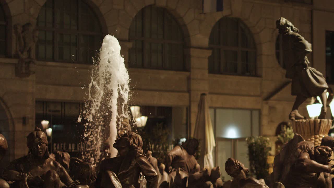 Bronze fountain statue sprays water upward in illuminated old town square, evening urban ambiance