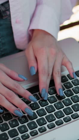 primer plano de manos con uñas pulidas escribiendo en el teclado de la computadora portátil al aire libre, sentado en un banco con una vista parcial de otra persona al lado, los detalles incluyen manicure elegante y trabajo activo
