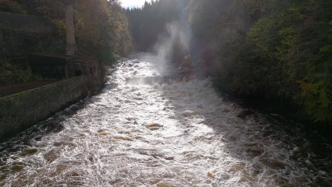슬로우 모션 드론 영상이 빠르게 흐르는 강과 오래된 건물로 둘러싸인 폭포 아래로 천천히 후퇴합니다.
