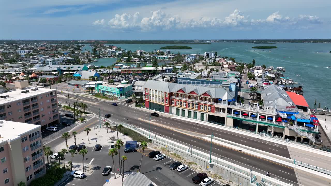 Aerial approaching shot of Madeira Beach with Johns Pass and driving cars in Florida. Colored buildings and palm trees along bay water.