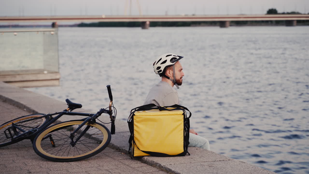 доставщик еды отдыхает рядом со своим терморюкзаком на закате на берегу реки и смотрит на свои смарт-часы в ожидании следующего заказа