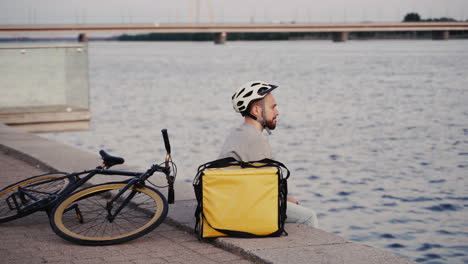 el repartidor de alimentos descansa junto a su mochila térmica al atardecer en la orilla de un río y echa un vistazo a su reloj inteligente esperando el próximo pedido