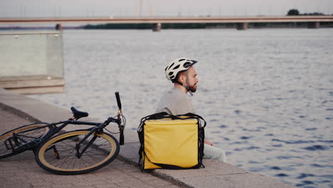 food delivery guy rests next to his thermal backpack at sunset on the shore of a river and takes a look at his smartwatch waiting for the next order