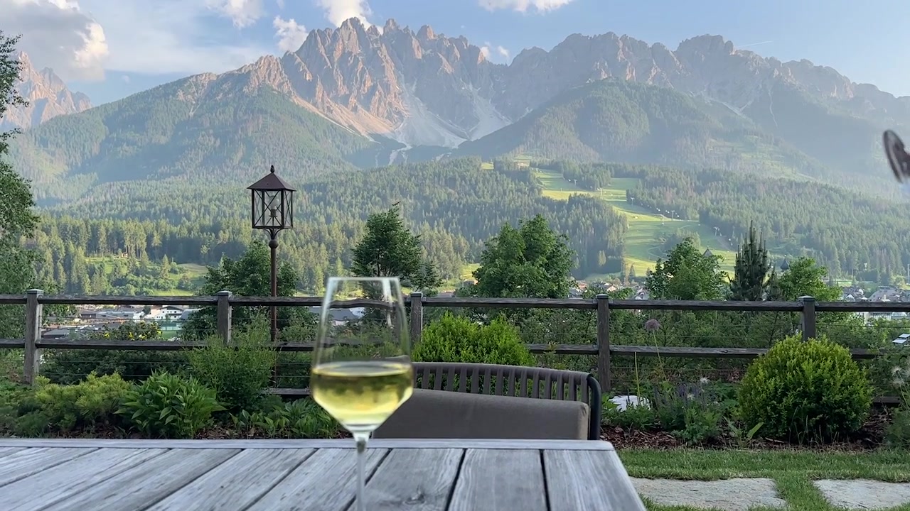 Elegantní lifestylový záběr dvou sklenic na víno zasazených do úchvatných panoramatických horských výhledů Dolomit v Jižním Tyrolsku – ruka sahá pro sklenici v tomto filmovém cestovatelském okamžiku.
