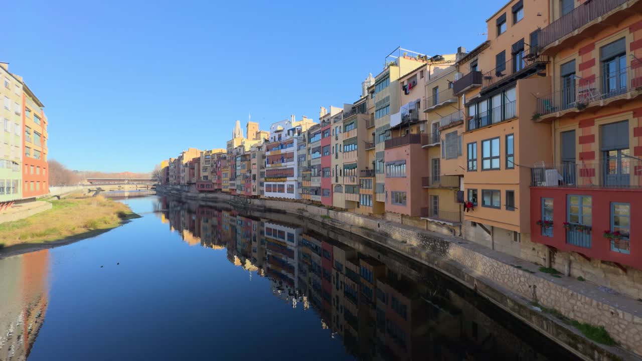 Brightly painted buildings line the tranquil riverbanks of girona, reflecting in the calm waters below a clear blue sky