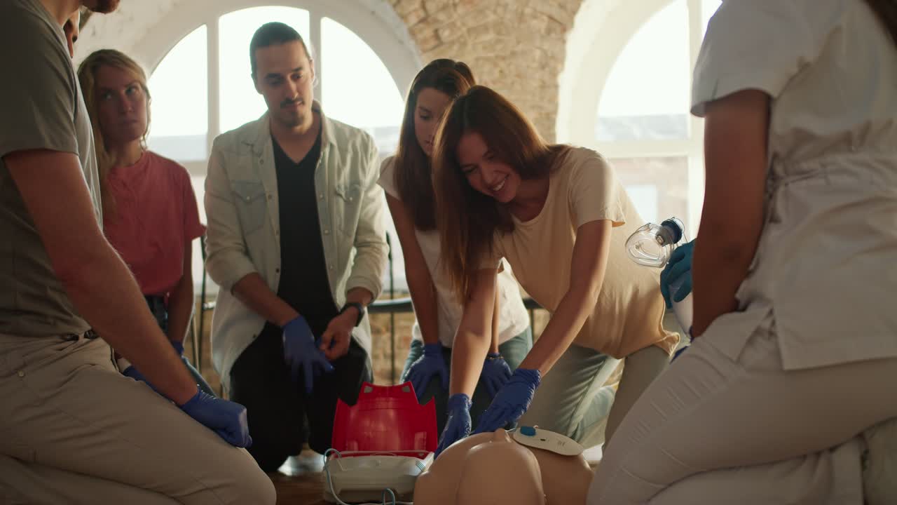 Close-up shooting: A girl in a white T-shirt learns to place medical electrical sensors on a mannequin's body under the instructions of a professional doctor, a girl in a white uniform, and a Black male doctor in a blue uniform, against a backdrop of white brick walls and large windows. A first aid 