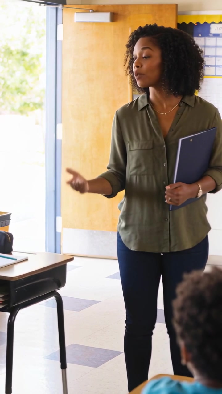Vertical video: Leaning forward, teacher in olive reaching, asking reply with blue folder in class