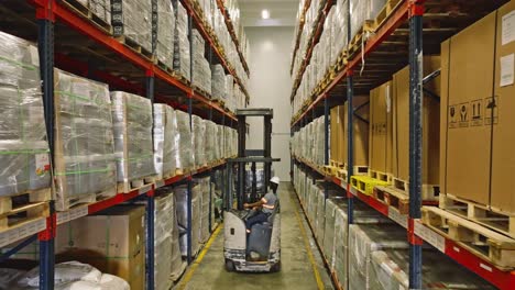 unrecognizable worker driving forklift between tall shelves of warehouse