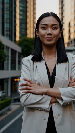 professional asian businesswoman confidently standing with arms crossed, radiating success in bustling urban street environment, embodying corporate leadership and modern career ambition