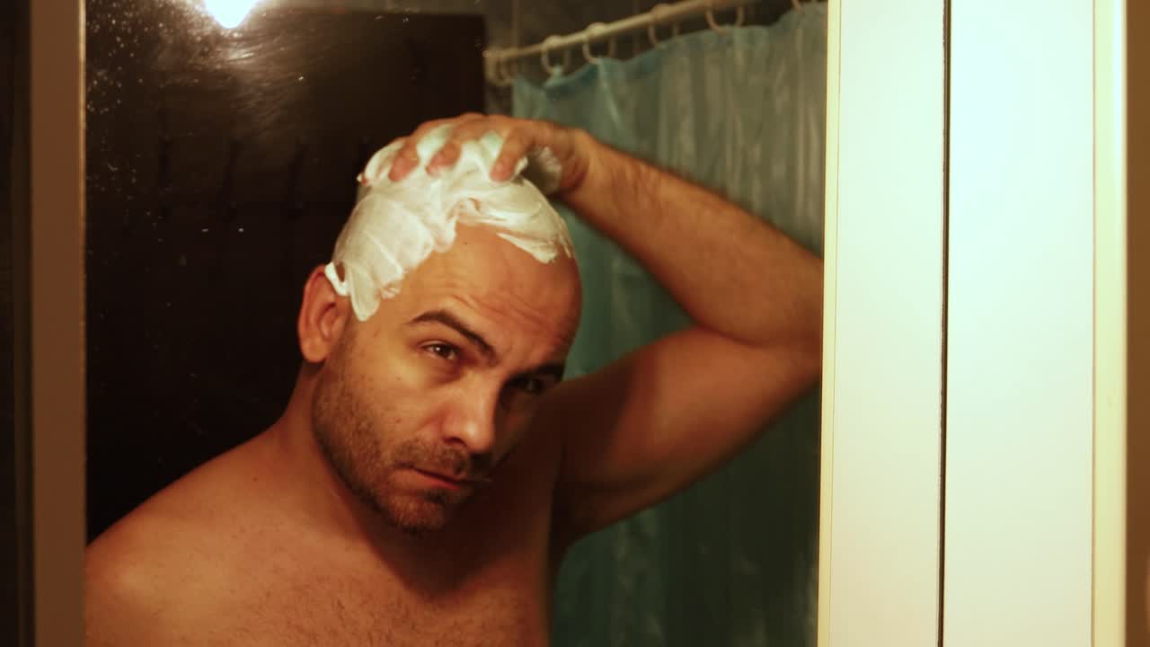 una foto de mal humor de un hombre barbudo calvo, aplicando espuma de afeitar a su cabeza, preparándola para un afeitado, frente al espejo del baño