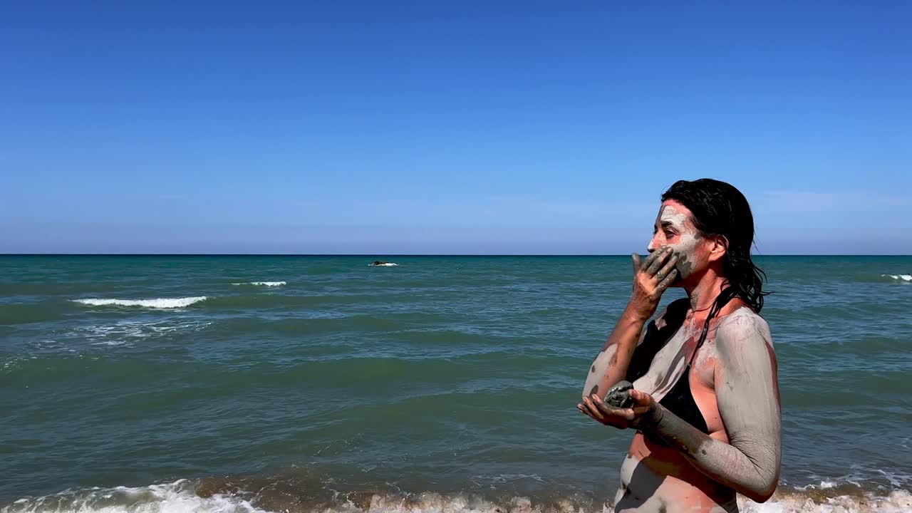 bella mujer caucásica en bikini negro esparce barro cosmético o arcilla en la piel de su cuerpo