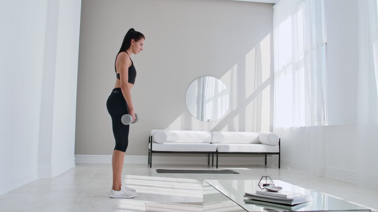The beautiful female performs leans forward with dumbbells in her hands in the white interior of the living room. In slow motion to do exercises on the muscles of the thighs