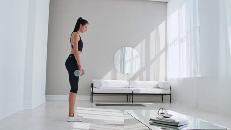 the beautiful female performs leans forward with dumbbells in her hands in the white interior of the living room. in slow motion to do exercises on the muscles of the thighs