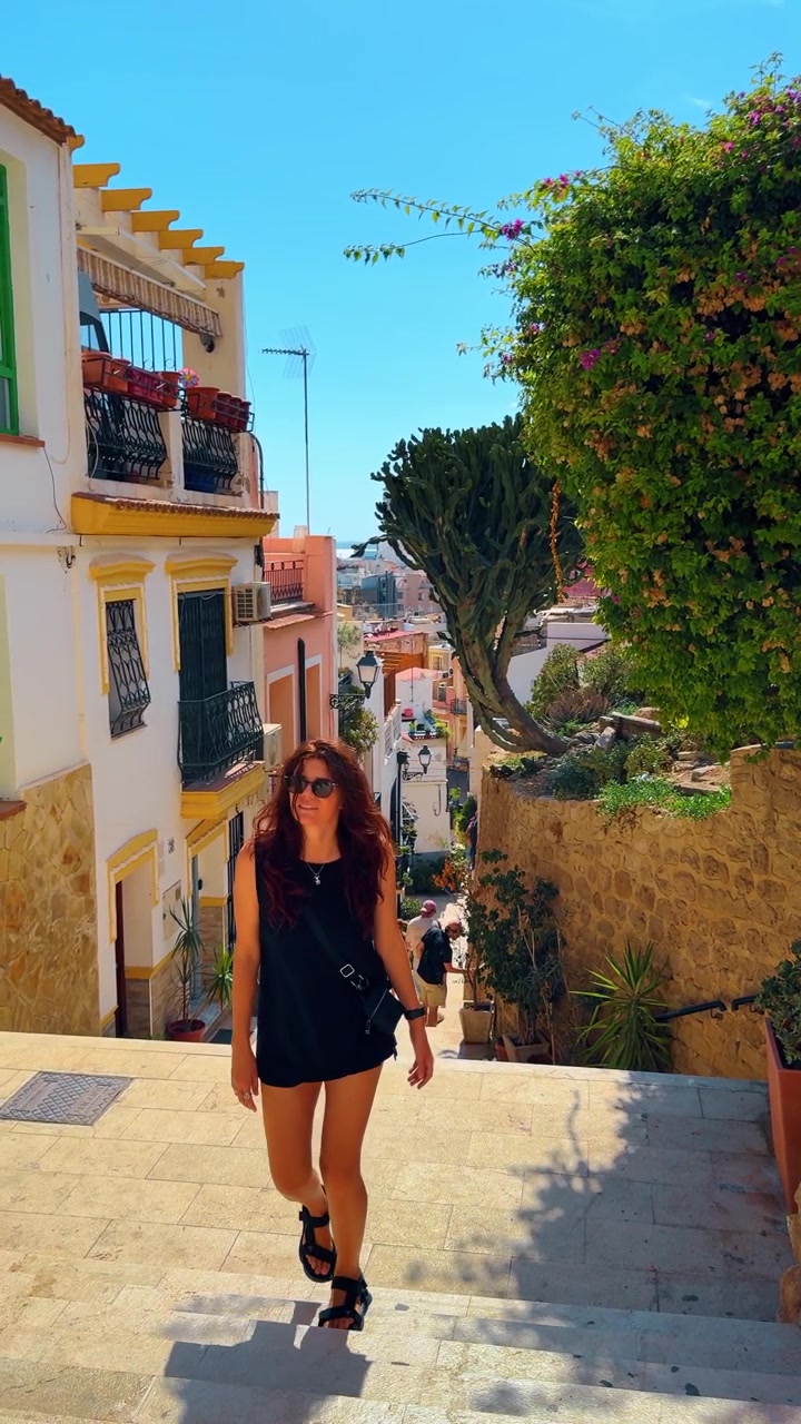 Alicante, Spanien, 8. august 2025: Kvinde går op ad stejle stentrappe i en gade i Alicante. Vertikal visning af en smal opadgående gade med farverige bygninger, trapper og en kvinde, der går mod kameraet.