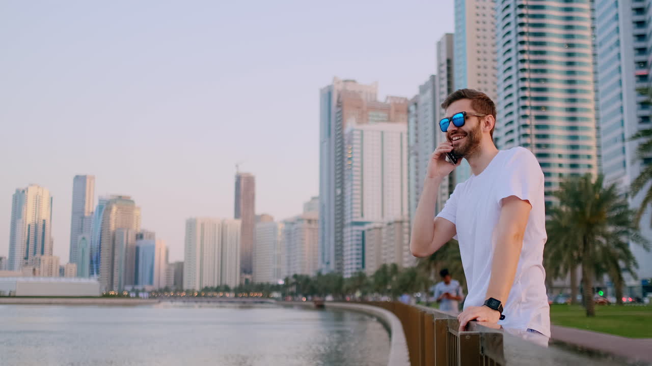 Young male tourist in sunglasses and bristles hipster in white t-shirt standing on the waterfront in the background of modern city talking on the phone and listening to the answer