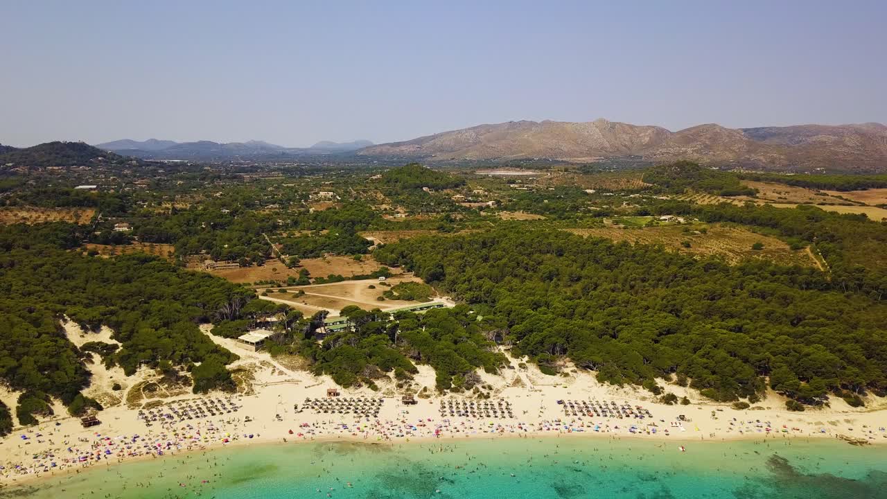 playa de cala agulla con bañistas de sol, rodeada de exuberante vegetación y montañas en el fondo, al mediodía, vista aérea
