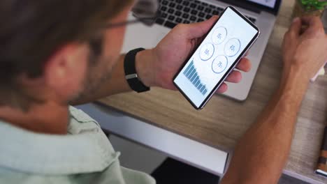 Caucasian-man-sitting-at-desk-using-smartphone-with-statistics-on-screen